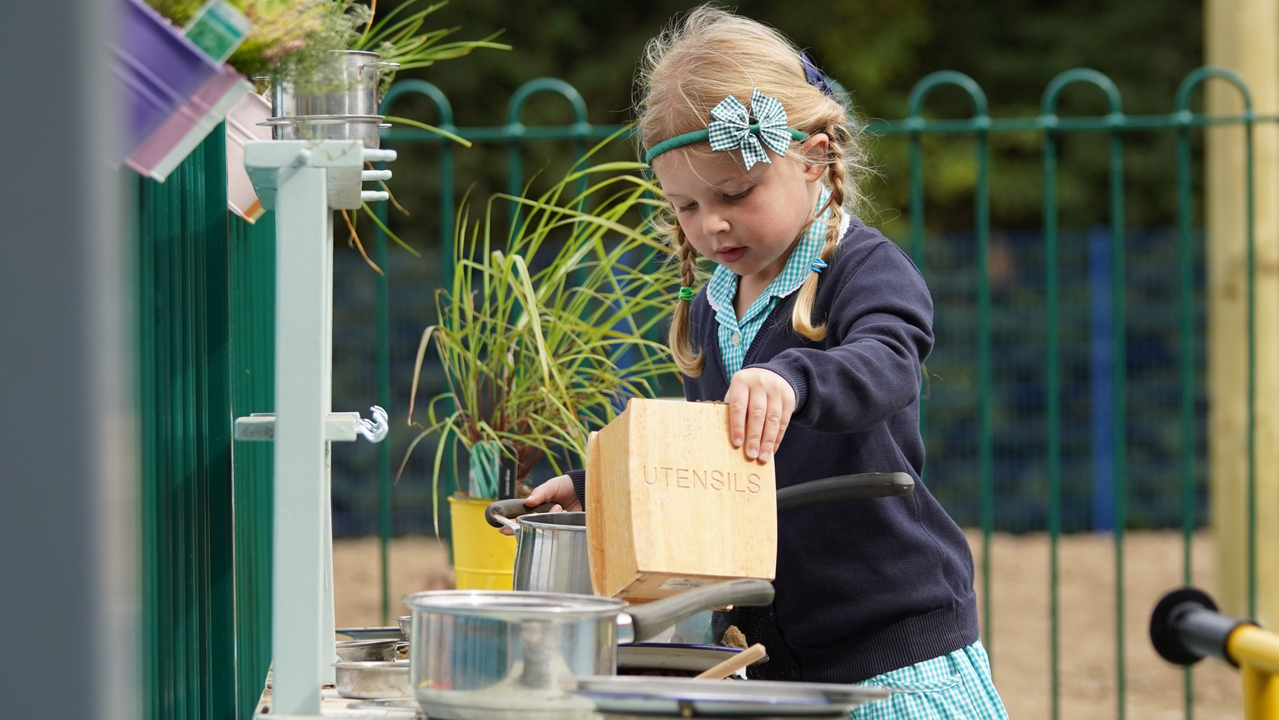 A young girl with blonde hair is seen interacting with some cooking utensils in an outdoor play area on the academy grounds.