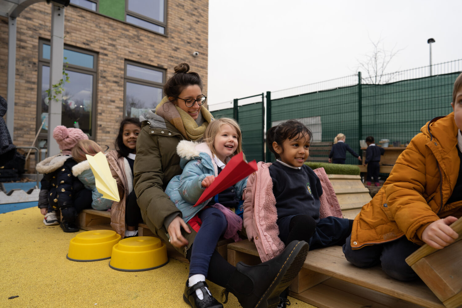 A small group of pupils are seen sat together along a wooden bench, wearing their winter coats, under the supervision of a female member of staff.