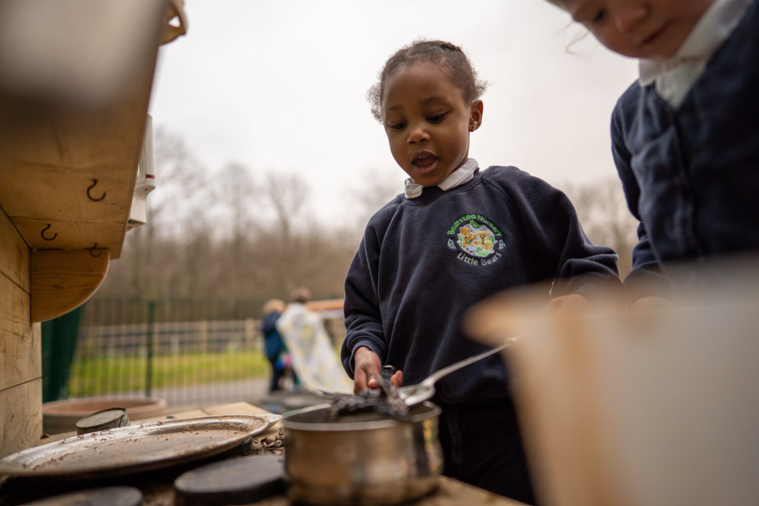Two young Nursery girls are pictured working with cooking utensils to mix a cake mixture. They are outdoors in a play area on the academy grounds.