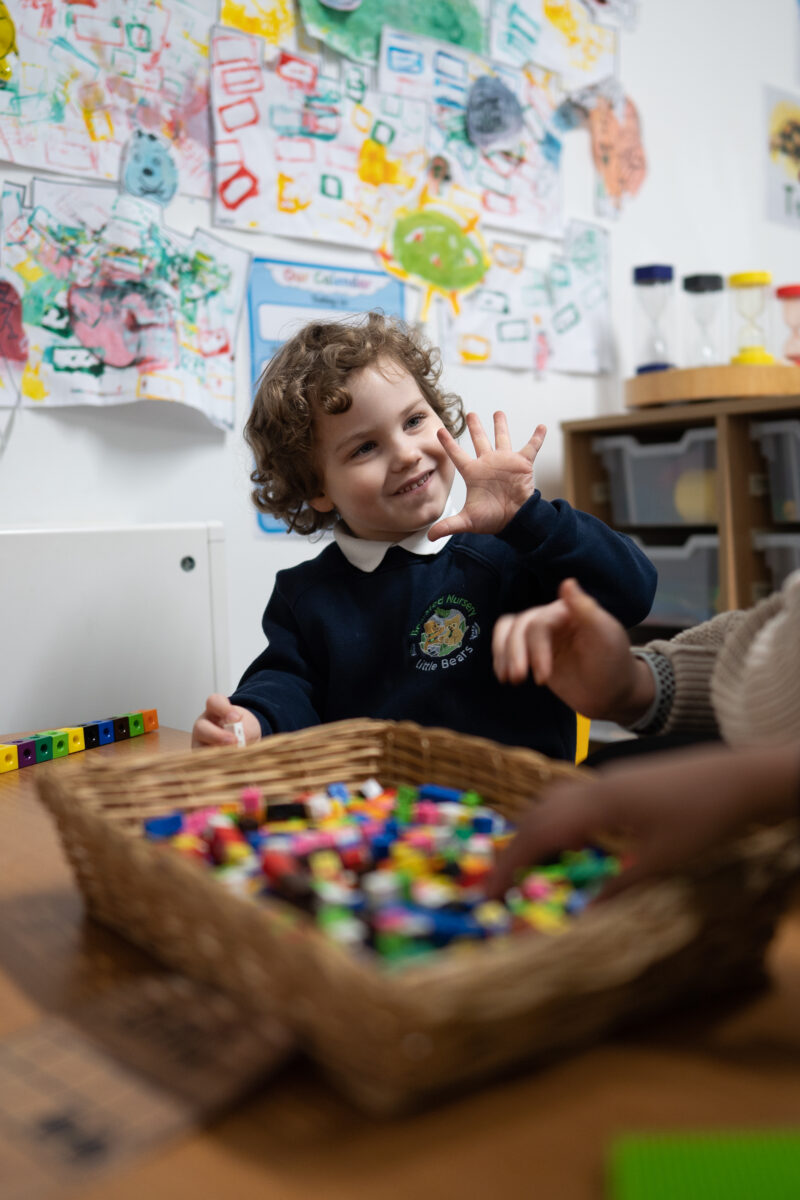 A young boy from Nursery with long, curly hair is seen wearing his uniform and playing with some counting cubes in class.
