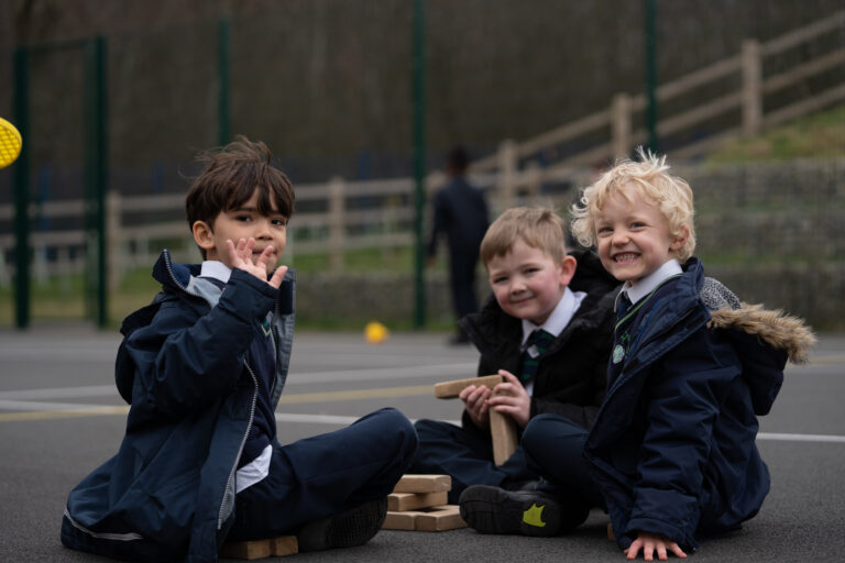 Three young boys are shown sitting on the ground and playing with wooden blocks together in an outdoor area on the academy grounds. All three are wearing their winter coats.