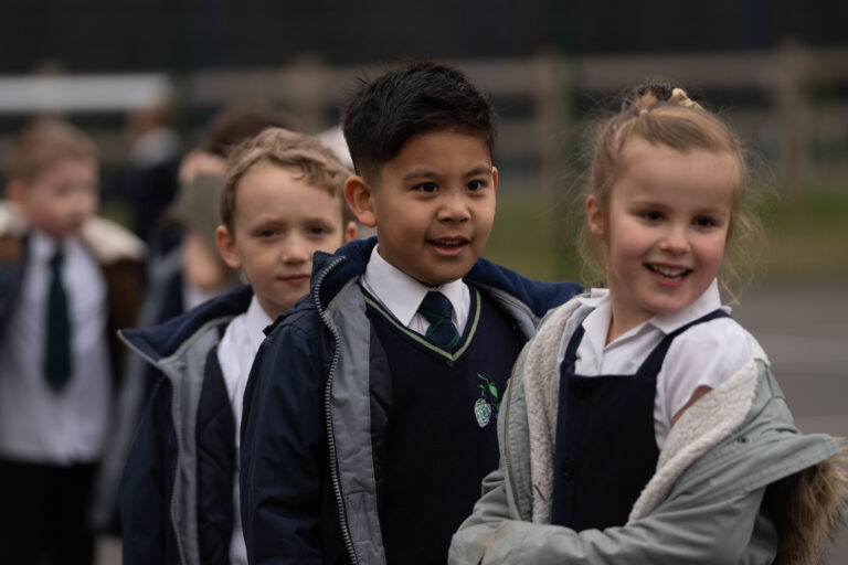 A group of three pupils, two boys and one girl, are seen standing outdoors on the academy grounds and smiling up ahead at something in front of them.